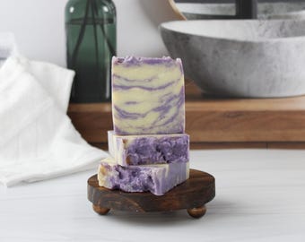 May include: A stack of three rectangular bars of soap with a marbled pattern of cream and purple colors. The soap bars are resting on a small, round, dark brown wooden pedestal stand with three short legs, showcasing a textured, uneven surface.