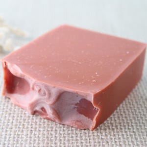 May include: A rectangular bar of soap in a muted dusty rose color. The soap features a smooth, flat top surface and a textured, swirled pattern along one of its sides, showing a slightly lighter shade of pink within the marbled design.
