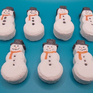 May include: Seven white snowman-shaped bath bombs arranged in two rows. Each snowman features a black top hat, an orange scarf, and small black dots representing eyes, a nose, and buttons on the torso. The figures have a textured, porous surface.