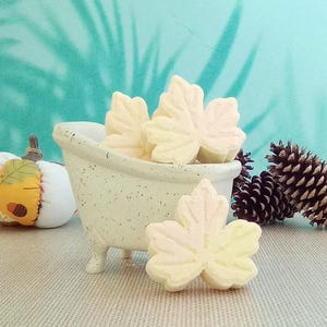 May include: A miniature cream-colored bathtub with speckled texture contains several pale yellow maple leaf-shaped bath bombs. One additional leaf-shaped bath bomb rests in front of the tub, accompanied by a fabric pumpkin and two pinecones.