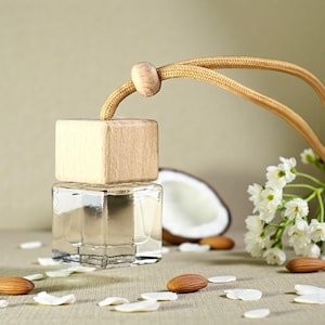 May include: A square glass bottle with a light-colored wooden cap and a tan cord for hanging. The bottle contains a clear liquid and is positioned near a halved coconut, white flower blossoms, and scattered almond nuts on a neutral surface.