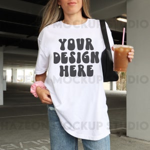 May include: A white oversized t-shirt with the text "YOUR DESIGN HERE" in black. The shirt is being worn by a person in a casual setting.