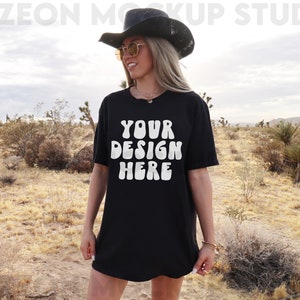 May include: A black oversized t-shirt with the text "YOUR DESIGN HERE" in white, worn by a person in a desert setting.