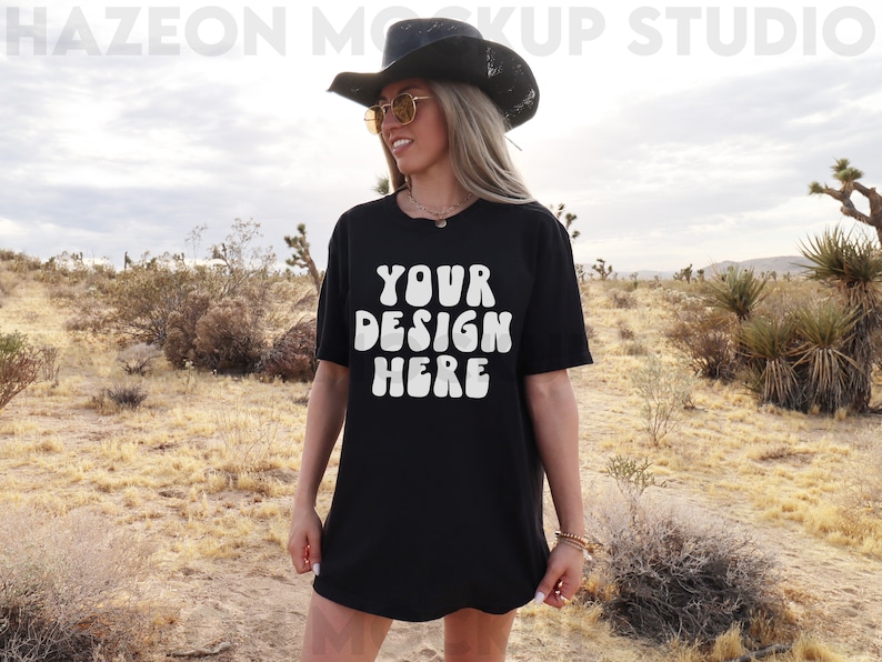 May include: A black oversized t-shirt with the text "YOUR DESIGN HERE" in white, worn by a person in a desert setting.