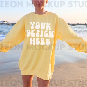 May include: A yellow sweatshirt with the text "YOUR DESIGN HERE" in white on the front. The sweatshirt is being worn by a person standing on a beach.