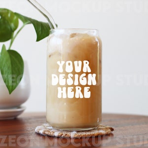 May include: A glass of iced coffee with a straw in a clear glass can with the text "YOUR DESIGN HERE" in white letters. The can is sitting on a woven coaster on a wooden table.