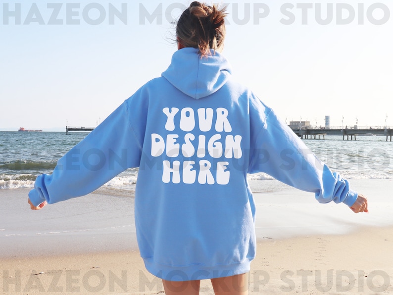 Peut inclure: Un sweat-shirt bleu clair avec le texte "YOUR DESIGN HERE" en blanc, en lettres de style r&eacute;tro. Le sweat-shirt est port&eacute; par une personne debout sur une plage de sable avec l'oc&eacute;an en arri&egrave;re-plan.