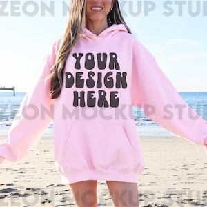 May include: A pink hoodie with the text "YOUR DESIGN HERE" in brown letters. The hoodie is being worn by a person standing on a beach.