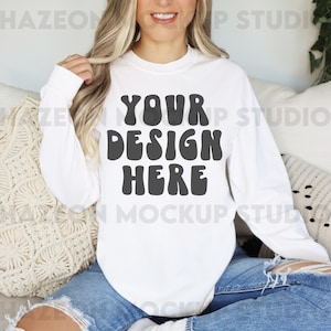 以下が含まれることがあります： 大胆なレトロフォントで「YOUR DESIGN HERE」の文字が入った白い長袖シャツ。カスタムデザインを展示するのに最適なモックアップです。モデルは、破れた青いジーンズとシャツを着ています。
