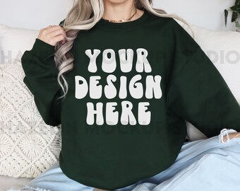 May include: A dark green sweatshirt with the text "YOUR DESIGN HERE" in white, retro-style lettering. The sweatshirt is being modeled by a person sitting on a white couch.