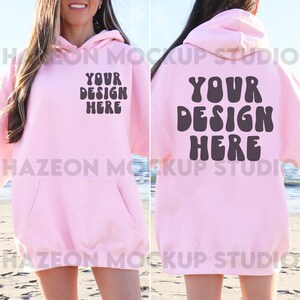 May include: A light pink hoodie with the text "YOUR DESIGN HERE" printed on the front and back. The hoodie has a front pocket and a hood. The image is a mockup of a hoodie on a person at the beach.