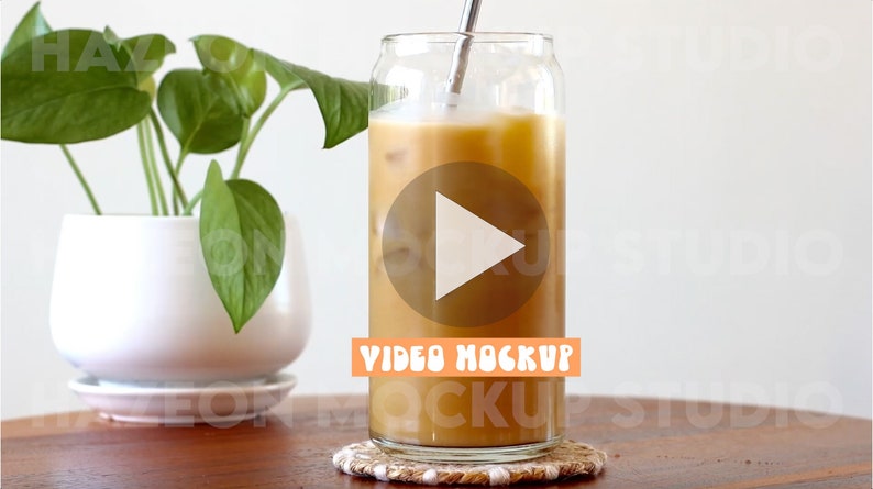 May include: A glass of iced coffee with a straw in a can-shaped glass. The coffee is light brown and there are ice cubes visible. The glass is sitting on a woven coaster. The text "VIDEO MOCKUP" is visible on the glass.