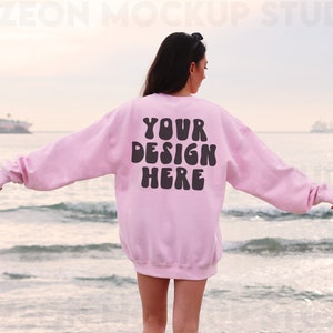 May include: A pink sweatshirt with the text "YOUR DESIGN HERE" in black letters on the back. The sweatshirt is being worn by a person standing on a beach.