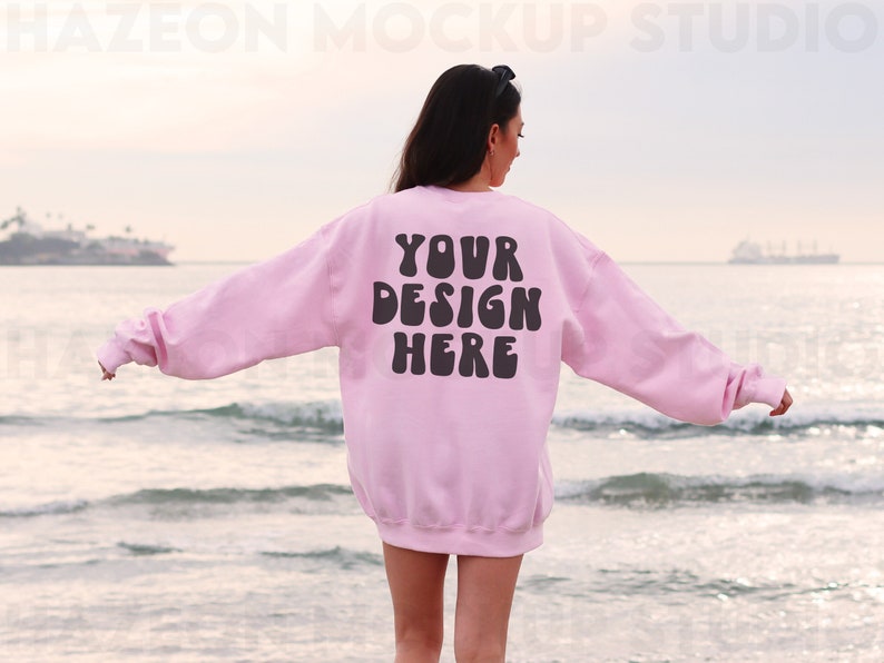 May include: A pink sweatshirt with the text "YOUR DESIGN HERE" in black letters on the back. The sweatshirt is being worn by a person standing on a beach.