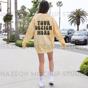 Może przedstawiać: Żółta bluza z tie-dye z napisem "YOUR DESIGN HERE" w brązowych literach na plecach. Bluzę nosi osoba idąca chodnikiem.