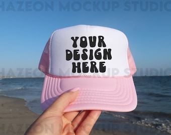 Op de afbeelding: Een roze en witte truckercap met een mesh achterkant. De voorkant van de cap heeft de tekst "YOUR DESIGN HERE" in zwarte, retro-stijl letters.