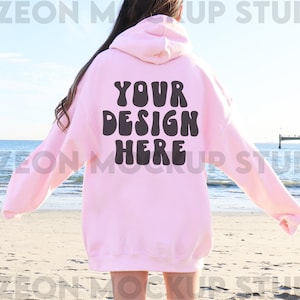 May include: A pink hoodie with the text "YOUR DESIGN HERE" in black, retro-style lettering. The hoodie is being worn by a person standing on a beach.