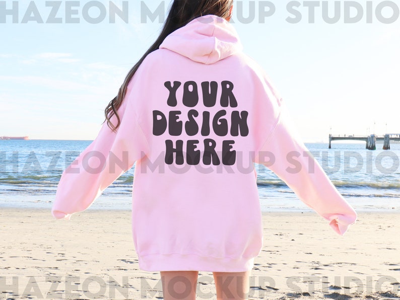 May include: A pink hoodie with the text "YOUR DESIGN HERE" in black, retro-style lettering. The hoodie is being worn by a person standing on a beach.