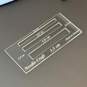 K&ouml;nnte beinhalten: Eine durchsichtige, rechteckige Acryl-Schablone f&uuml;r Handarbeiten mit vier rechteckigen Aussparungen, die mit 0,5 cm, 0,7 cm, 1,0 cm und 1,5 cm beschriftet sind. Das Werkzeug zeigt zudem den Text Needle Craft, 2,0 cm, 3,0 cm und Made in Ukraine.