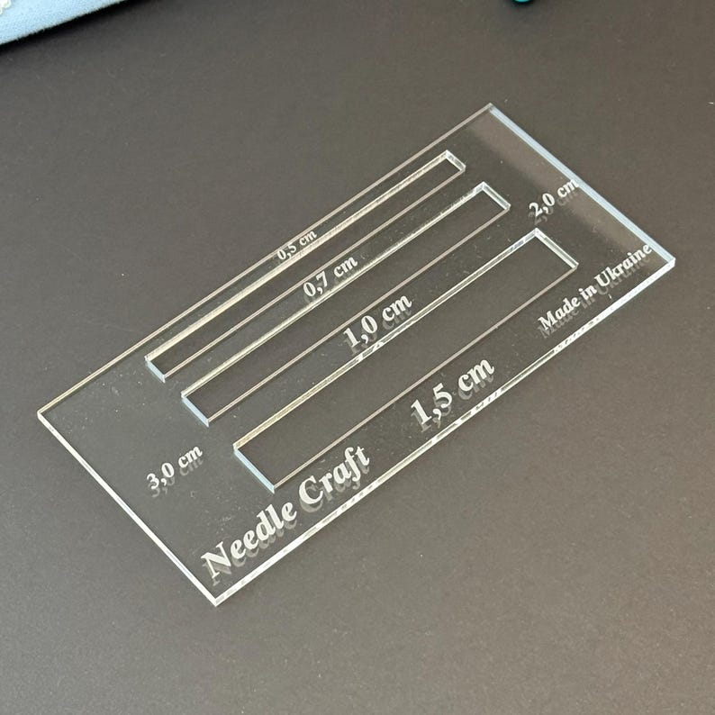 K&ouml;nnte beinhalten: Eine durchsichtige, rechteckige Acryl-Schablone f&uuml;r Handarbeiten mit vier rechteckigen Aussparungen, die mit 0,5 cm, 0,7 cm, 1,0 cm und 1,5 cm beschriftet sind. Das Werkzeug zeigt zudem den Text Needle Craft, 2,0 cm, 3,0 cm und Made in Ukraine.
