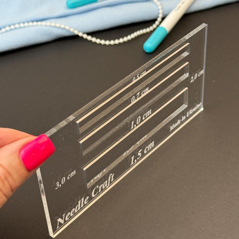 K&ouml;nnte beinhalten: Eine transparente Acryl-Schablone f&uuml;r Handarbeiten mit mehreren rechteckigen Aussparungen, die mit Ma&szlig;en in Zentimetern beschriftet sind, darunter 0,5 cm, 0,7 cm, 1,0 cm, 1,5 cm, 2,0 cm und 3,0 cm, sowie dem Aufdruck Needle Craft und Made in Ukraine.