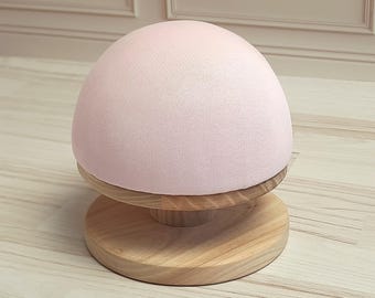 May include: A pink fabric covered dome head form on a wooden base. The base is round and has a slight lip around the edge.