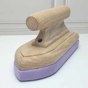 Può includere: Un asse da stiro in legno con una copertura imbottita viola. L'asse ha una forma curva e una maniglia rialzata.