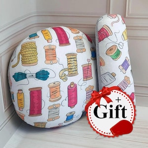 May include: Two round throw pillows with a white background and colourful spools of thread, yarn, and needles printed on them. The larger pillow has the text "+ Gift" printed on it.