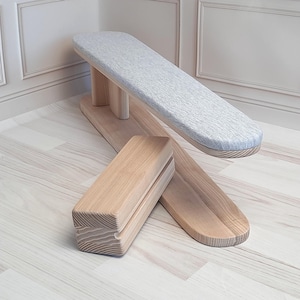 Può includere: Un asse da stiro in legno con una superficie imbottita grigia e un blocco di legno per premere le cuciture. L'asse è in legno chiaro e ha una finitura naturale.