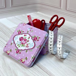 May include: A pink metal tin with a floral pattern and white polka dots. The tin is open and contains red scissors, a red measuring tape, and a red pen. The tin is sitting on a light wood surface.