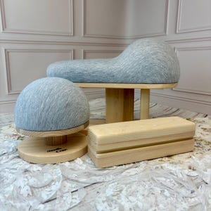May include: A set of three wooden sewing tools with light grey fabric covers. The set includes a dome-shaped tailor's ham, a curved pressing cushion on a wooden stand, and a rectangular wooden pressing block with the text Needle Craft printed.