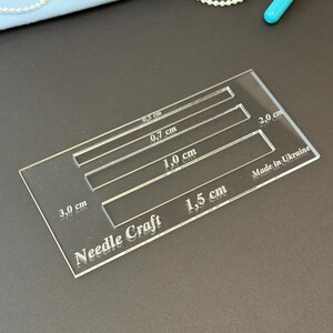 K&ouml;nnte beinhalten: Eine durchsichtige, rechteckige Acryl-Schablone f&uuml;r Handarbeiten mit vier horizontalen Aussparungen, beschriftet mit 0,5 cm, 0,7 cm, 1,0 cm und 1,5 cm. Das Werkzeug tr&auml;gt die Gravuren Needle Craft, 3,0 cm, 2,0 cm und Made in Ukraine.