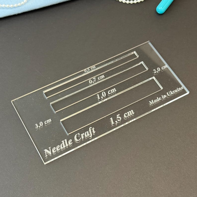 K&ouml;nnte beinhalten: Eine durchsichtige, rechteckige Acryl-Schablone f&uuml;r Handarbeiten mit vier horizontalen Aussparungen, beschriftet mit 0,5 cm, 0,7 cm, 1,0 cm und 1,5 cm. Das Werkzeug tr&auml;gt die Gravuren Needle Craft, 3,0 cm, 2,0 cm und Made in Ukraine.