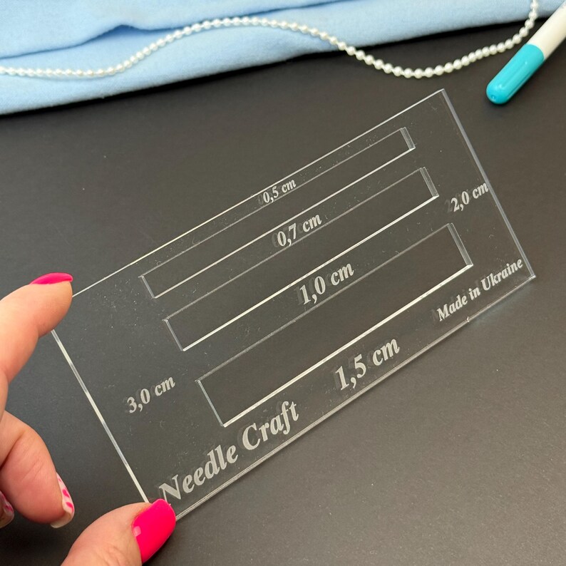 K&ouml;nnte beinhalten: Eine transparente, rechteckige Acryl-Schablone f&uuml;r Handarbeiten mit vier horizontalen Aussparungen, die mit metrischen Ma&szlig;en von 0,5 cm, 0,7 cm, 1,0 cm und 1,5 cm versehen sind. Das Werkzeug tr&auml;gt die Aufschrift Needle Craft und Made in Ukraine.