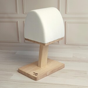 May include: A white, padded hat stand with a wooden base and post. The base is rectangular, and the post is square. The top is a rounded, padded surface. The words "Needle Craft" are engraved on the base.