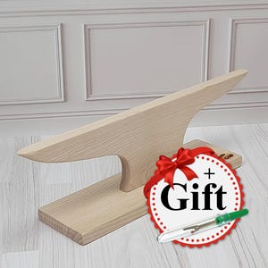 Può includere: Incudine in legno con una base larga e piatta e una parte superiore appuntita rialzata. L'incudine è realizzata in legno chiaro e ha una finitura naturale. Un fiocco rosso e un cerchio bianco con il testo "Regalo" sono in primo piano.