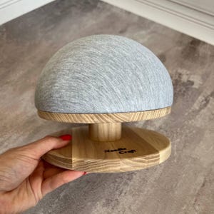 May include: A grey fabric covered dome sits on a wooden base with a circular wooden platform. The base has the words "Needle Craft" written on it.