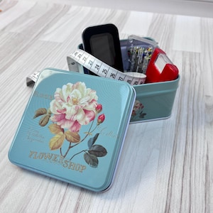 May include: A light blue metal sewing box with a floral design and the text "FLOWER SHOP". The box contains a measuring tape, a small black device, and sewing supplies. The box is open, revealing its contents.