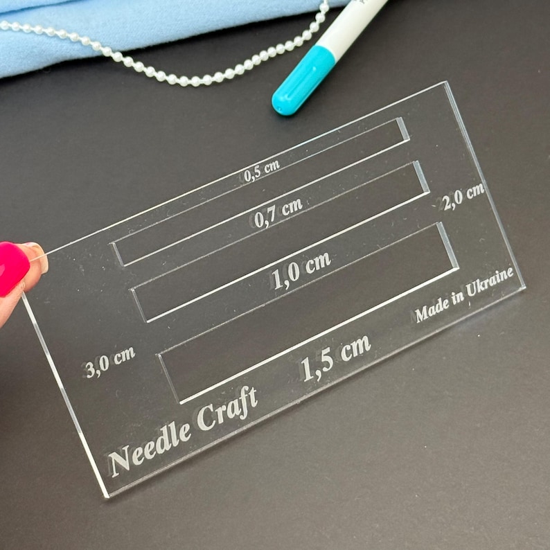K&ouml;nnte beinhalten: Eine durchsichtige, rechteckige Acryl-Schablone f&uuml;r Handarbeiten mit vier rechteckigen Aussparungen, die mit 0,5 cm, 0,7 cm, 1,0 cm und 1,5 cm beschriftet sind. Das Werkzeug tr&auml;gt die Gravuren Needle Craft, 2,0 cm, 3,0 cm und Made in Ukraine.