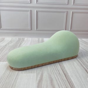 Pode incluir: Um almofadinha de alfinetes de costura coberto com tecido verde claro com uma base de madeira. O almofadinha de alfinetes tem a forma de um oval longo e curvo.