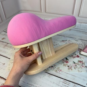 Op de afbeelding: Een roze opgevulde kleermakersham met een houten basis. De ham heeft een levendige roze kleur en wordt ondersteund door een lichtgekleurd houten frame. De basis is licht van kleur.