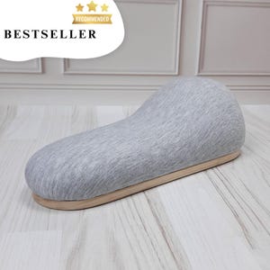May include: A gray fabric and wood footrest shaped like a crescent moon. The footrest is on a white wood floor.