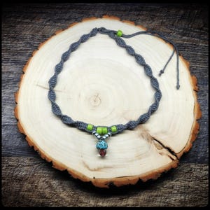 May include: A gray macrame necklace with green beads and a mushroom pendant. The pendant has a blue and white design with a red bead at the bottom. The necklace is displayed on a wooden slice.
