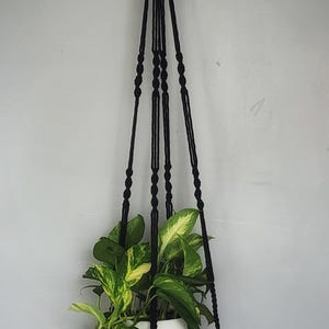 May include: A black macrame plant hanger featuring a circular woven rattan shelf suspended by knotted cords. The hanger holds a white ceramic pot containing a green and yellow variegated pothos plant, with a light wood ring at the top for hanging.