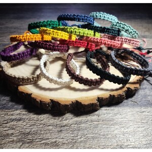 May include: A collection of macrame bracelets in various colors, including blue, green, yellow, and purple, are displayed on a wooden slab. The bracelets are handmade and feature a woven design, showcasing a range of hues and textures.