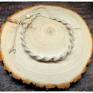 May include: A handmade, beige macrame necklace with a spiral design. The necklace is made of natural fiber and features a wooden bead closure. The necklace is displayed on a wooden slice with a natural wood grain.