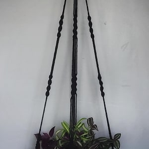 May include: A black macrame plant hanger featuring a circular wooden tray base with handles. The hanger is suspended from a light-colored wooden ring and includes a decorative black tassel at the bottom, holding a plant with purple and green leaves.