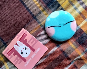 May include: Two pins on a plaid fabric background. One pin is a light blue circle with a white and pink face. The other pin is a pink square with a white rabbit in a red torii gate. The text "FLOOFY BUN TIME" is on the pink square.