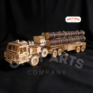 May include: A detailed wooden model of a military truck with multiple wheels and cylindrical structures. The model is made of light brown wood and features intricate cut-out details. The text "ONLY ARTS COMPANY" is visible.