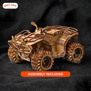 May include: A wooden ATV model with intricate laser-cut details. The model features large, textured wheels and a detailed frame. The text "ASSEMBLY INCLUDED" is displayed on an orange banner. The Only Arts Company logo is in the upper left corner.